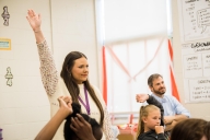 A teacher trainee enrolled in an apprenticeship program at Reach University stands in front of an elementary school classroom, her hand raised as if to get students' attention. In the background, Reach University president Joe E. Ross is seated.
